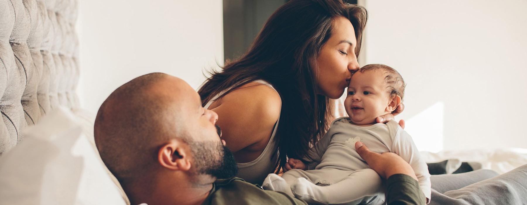 mother kisses her infant while father lies in bed and holds him